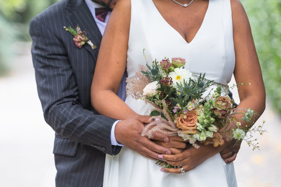 A bride wearing a form-fitting white wedding gown with intricate lace embroidery and a row of buttons down the front, holding a bouquet of fresh flowers. The bouquet features white calla lilies, pink 
