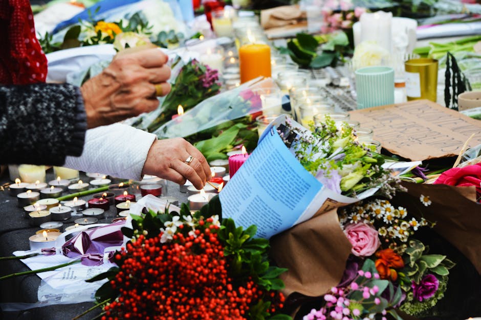 A close-up view of a floral tribute consisting of vibrant red berries, pink roses, white daisies, and green foliage arranged on a table covered with numerous lit tealight candles in small white holder