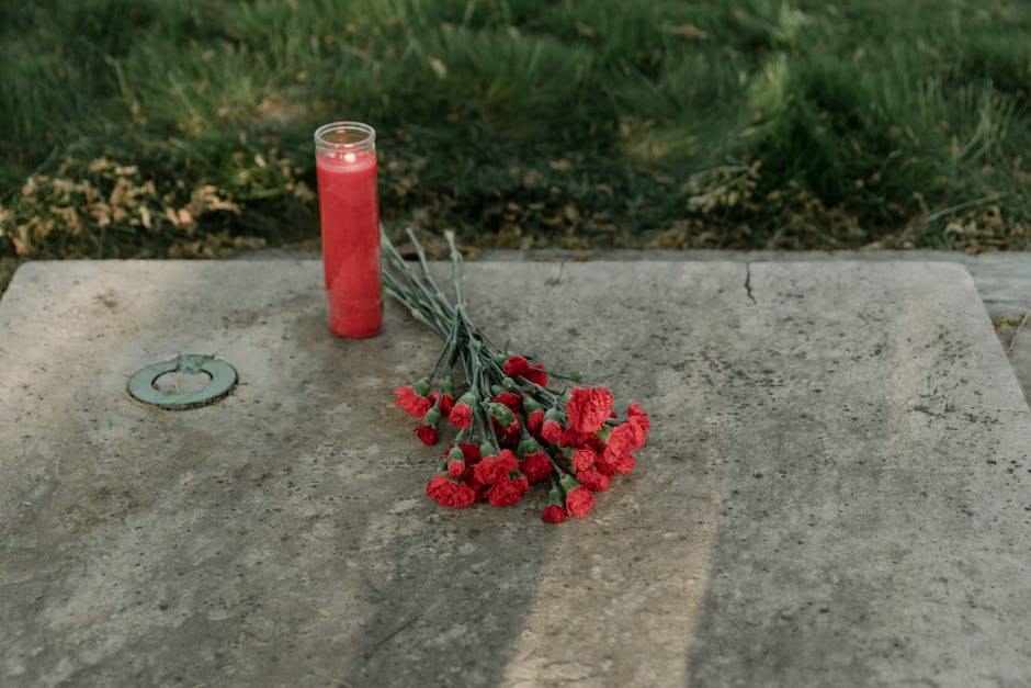 A bouquet of red carnations with green stems and leaves, arranged casually on a weathered, light gray concrete surface. Next to the flowers is a tall, cylindrical red candle, partially melted, with a 