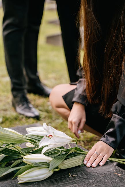 A person with long brown hair, wearing a black jacket and skirt, is kneeling at a gravesite, gently resting their hand on a bouquet of white lilies with green leaves that is placed on the grave stone.