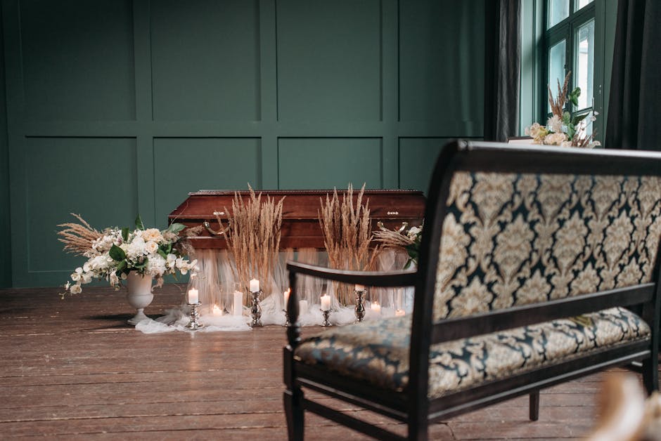 An elegant memorial setup with a closed wooden casket surrounded by floral arrangements and decorative candles inside a room with dark green paneled walls and large windows. To the left of the casket,