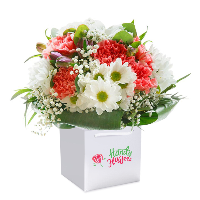 A radiant floral display, carefully composed to capture the essence of Golders Green, stands poised in a luminous, white square gift bag, set gracefully against a pure white backdrop. The bouquet features a lush blend of peach and pink-hued carnations, each bloom's ruffled petals catching the light and radiating warmth akin to a midsummer stroll along Finchley Road. Nestled between the larger blooms, crisp white daisies with central orbs of lime-green bring brightness and pop, reminiscent of wildflower patches found in Golders Hill Park. Lacing through the bouquet, delicate sprigs of baby's breath add an airy, cloudlike texture, while on the left a handful of buds tinged with purple-pink hues flirt with the eye, promising blooms yet to come and echoes of private gardens around Hampstead. Abundant, glossy green foliage creates a sturdy base, cradling the bouquet and forming a natural, elegant frame. The gift bag, complete with smooth ribbon handles, communicates readiness for delivery-be it for a special birthday, a noteworthy anniversary, or a heartfelt thank you to someone special. Subtle yet unmistakable, the attention to detail and freshness of each stem uplift the entire arrangement, making it the perfect emblem of quality and thoughtful local floristry for anyone in the Golders Green and NW11 area.