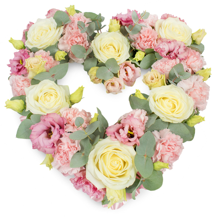 A softly coloured, heart-shaped wreath designed for funerals and memorials is presented against a pure white background, its lush fullness and gentle palette creating an atmosphere of calm reflection. The entire heart is formed from a tightly woven tapestry of blossoms, anchored by prominent cream and pale lemon roses scattered evenly across the design. Each rose appears plush and velvety, with thick petals curling from a compact centre into broader outer layers, catching subtle highlights and shadows that enhance their three-dimensional form. Between these roses, clusters of pastel pink carnations are tucked in, their many frilled petals building a billowing texture that gives the heart a sense of softness and depth. Richer, more saturated touches of mauve and magenta-pink lisianthus are dotted throughout the arrangement, their slightly ruffled, open blooms and occasional darker centres drawing the eye and adding a delicate complexity. Silvery-green eucalyptus leaves thread through the florals, looping in gentle arcs that both support and frame the heart, their cool, rounded foliage softening transitions between colours. Here and there, small yellow-green buds emerge, fresh and tightly closed, suggesting renewal even as the wreath speaks of farewell. The composition feels expertly balanced, neither too heavy nor too sparse, each flower placed with intention. It would be a fitting tribute in Golders Green, perhaps resting by a memorial plaque or placed atop a coffin during a service, ready to be carried to a final resting place. The heart motif symbolises enduring love and cherished memories, while the pastel tones and natural textures bring comfort and a quiet, dignified beauty to moments of grief.