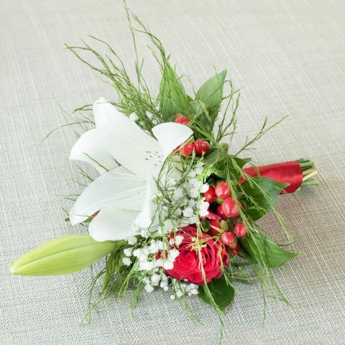 Viewed from above, a delicately crafted bridesmaid bouquet is thoughtfully showcased on a subtly textured light beige fabric, echoing the gentle elegance found in Golders Green's boutiques. The focal point, a white lily in full bloom, displays flawless petals gracefully spread open, distinguished by fine reddish-brown speckles and a striking cluster of yellow stamens. Just beneath, a luxuriant, deep red rose bursts with velvety petals, its intense hue contrasting beautifully and bringing a sense of passionate romance-much like the vibrant energy in the cafés of Finchley Road. Highlights of tiny white baby's breath create soft waves of texture and light, while glossy red hypericum berries cluster among the blooms, evoking festive gatherings and brightening the composition. On one side, a slender lily bud in light green suggests anticipation and the continual promise of new memories. An assortment of textured greenery, from airy feathery fronds to broad dark leaves, surrounds the bouquet, imbuing it with natural depth and movement. At the bouquet's base, slender stems are neatly joined and enveloped in a sleek red satin ribbon, drawing the crimson motif together. The whole piece is lit in diffused light, showing off the freshness and vibrancy of every element. This arrangement is an elegant, evocative choice for weddings, anniversaries, and cherished gifting moments in Golders Green.