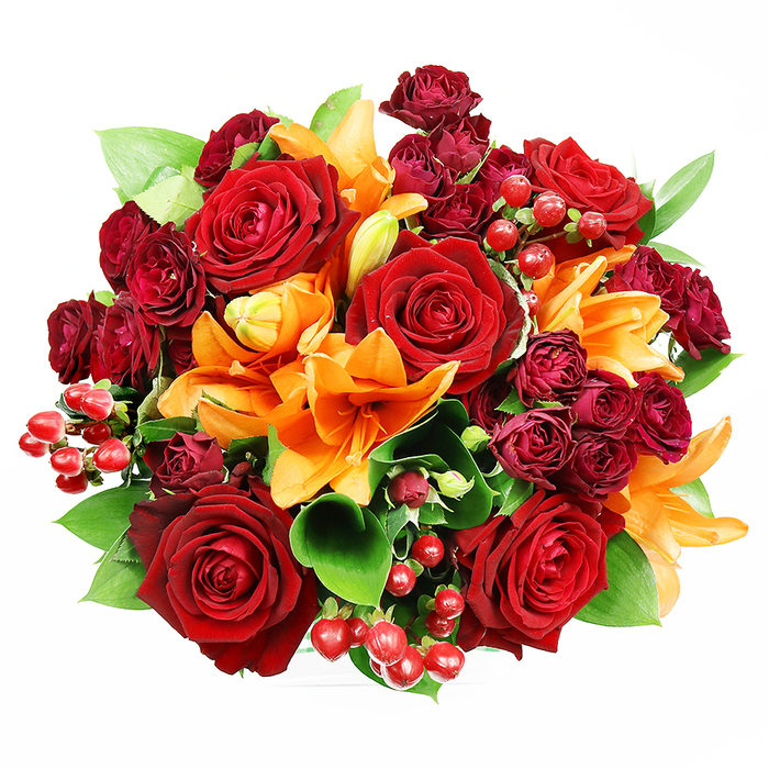 This lush, circular bouquet, photographed from above against a stark white ground, reads as an artful study in warm colour and tactile contrast. Dominant, fully opened deep red roses form bold focal points with their dense, velvety petals curling into a classic spiral-a visual shorthand for passion and devotion. Interspersed among them, bright orange Asiatic lilies introduce dramatic trumpet forms and exposed stamens that catch the eye; some lilies are at the peak of bloom, others in mid-unfurling stages that promise extended life and evolving texture. Numerous small, dark red spray roses cluster like scattered garnet cabochons, their compact blooms lending a luxurious, layered surface that deepens the overall composition. Glossy hypericum berries in clear ruby tones dot the arrangement, providing rhythmic highlights and a playful contrast of roundness against petal layers. Broad, glossy greenery-suggestive of ruscus or salal-emerges between blossoms to provide structure, freshness and a cool colour foil to the warm palette. The lighting is gentle and uniform, revealing the satiny sheens and delicate veining on each petal without harsh shadow, while the combined scent hints at spicy lily top notes resting on the warm, familiar perfume of newly cut roses. This bouquet feels perfectly suited for an autumn anniversary, a romantic declaration or a celebratory gift for a home near Temple Fortune or the shops on Golders Green Road, an arrangement that brings both visual theatre and comforting familiarity.