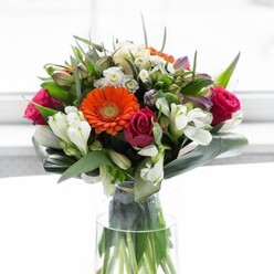 This radiant bouquet, handcrafted by Golders Green Flowers, is a true celebration of colour, texture, and local flair. Prominent are brilliant orange and peach gerbera daisies, their petals luminous beneath a wash of natural light, each bloom evoking the cheerful energy of early-morning strolls on Finchley Road. Beside them, magenta roses curl tightly, their richness balancing the arrangement with an unmistakable sense of romance. White alstroemeria and dainty spray roses brighten the palette, while clouds of lighter pink and petite white filler flowers create a sense of layered abundance. Glossy green leaves in various sizes add structure and contrast, with large, elegantly curved aspidistra fronds wrapping the base in a protective embrace and echoing the parkland tranquility found nearby. The bouquet sits in a clear glass vase-a nod to pure, understated elegance-allowing the vividly colored stems to shine through, grounded in their natural beauty. Illuminated by gentle, diffuse daylight that brings every petal and leaf into vibrant focus, this bouquet conjures feelings of hope and celebration: perfect for marking achievements, welcoming new chapters, or sending heartfelt wishes to friends and family in Golders Green.